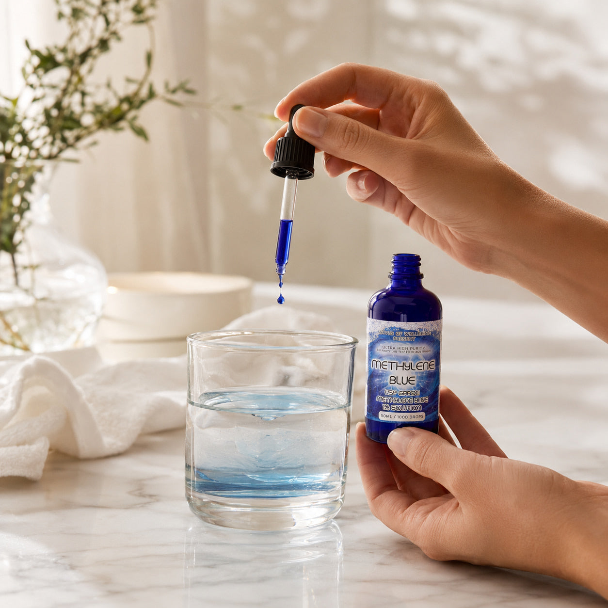 Hand holding a bottle of Methaqua Blue and dropping it into a glass of water on a marble surface.